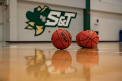 Basketballs sit on a glossy wooden gym floor, an S&T athletics logo can be seen in the background